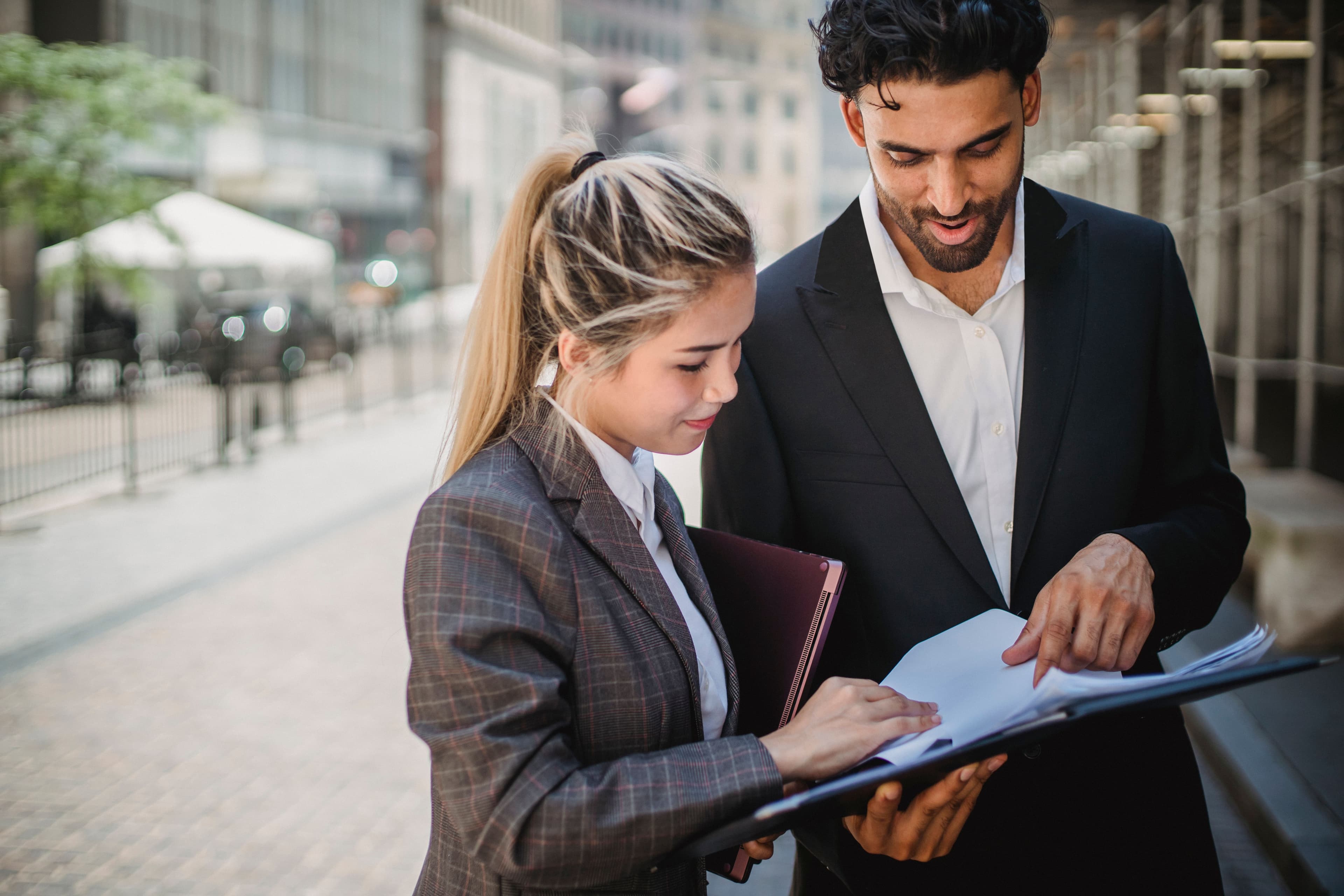 Two business colleagues standing on a city street, reviewing paperwork together in a folder.