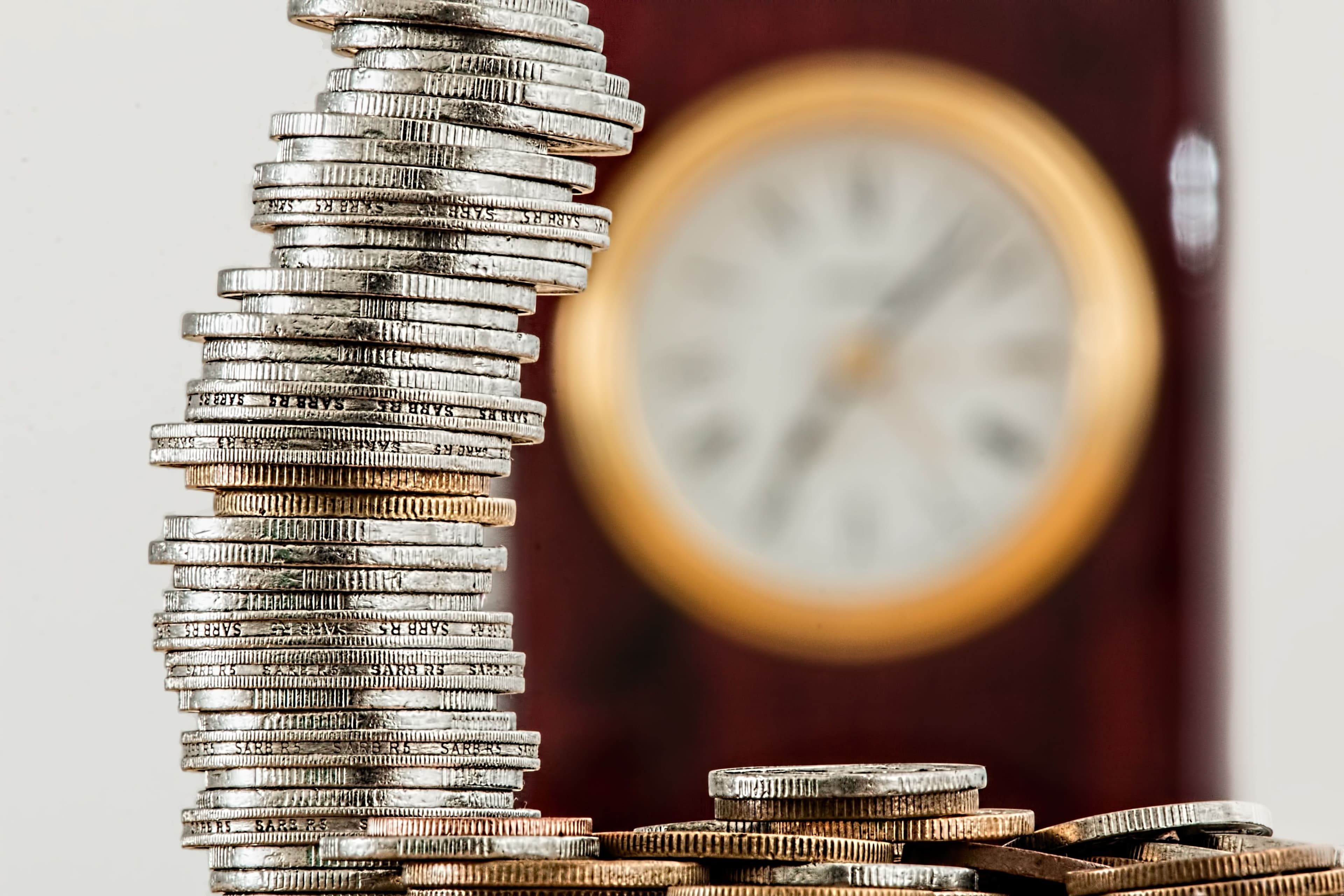 A tall stack of coins in front of a blurred clock face, suggesting the cost and timing of setting up a company.