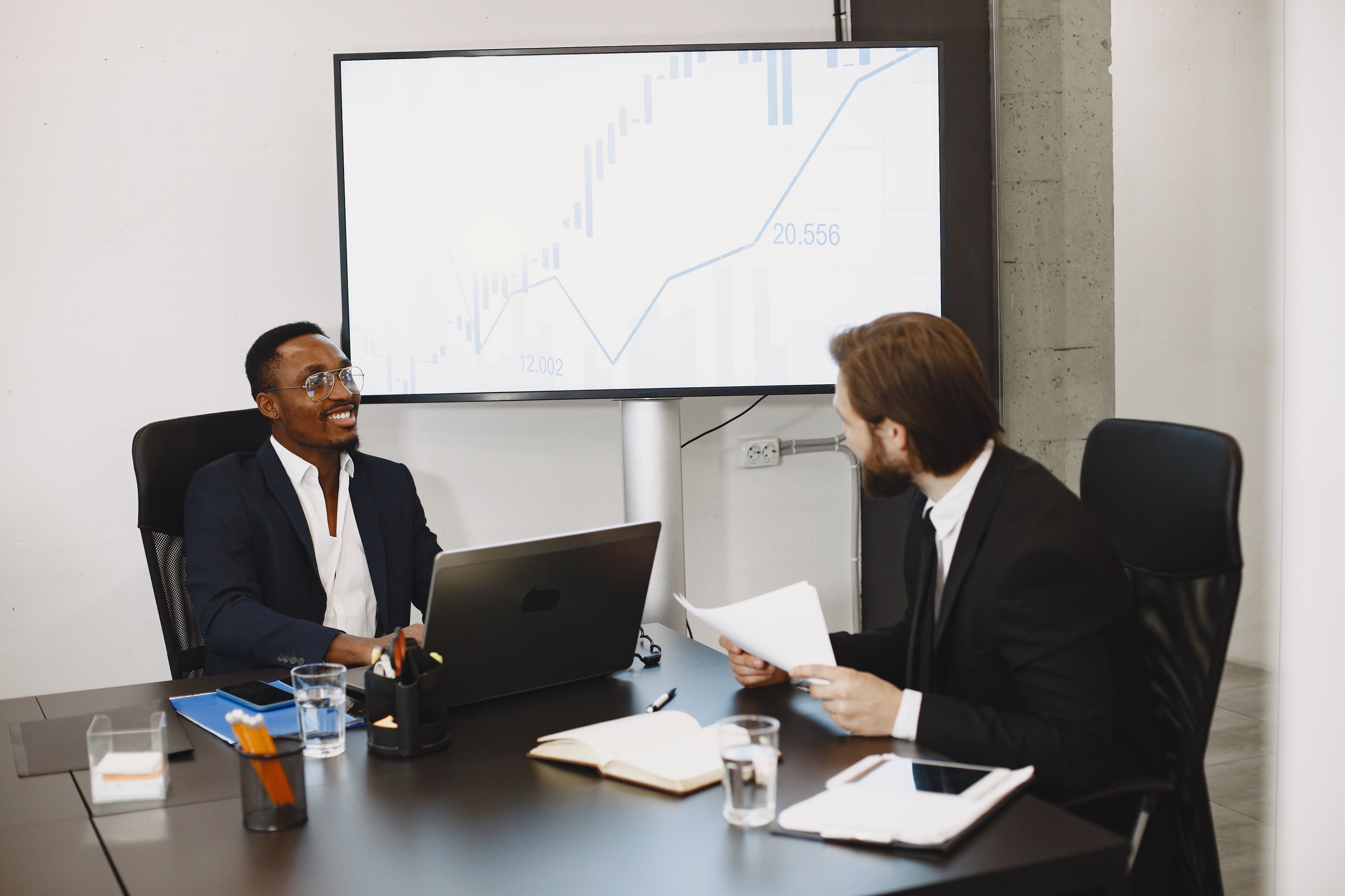 Two business owners talking across a desk with a growth chart displayed on a screen behind them.